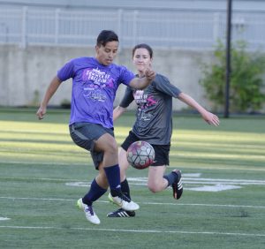 two people playing LGBT league soccer in a field outside during the day. both players have different-colored shirts that say "summer of freedom" and a sponsor message from Nellie's sports bar