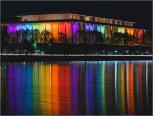 the Kennedy Center in Washington, DC, lit up rainbow; the photo is taken at night and the rainbow lights reflect on the Potomac River in the foreground